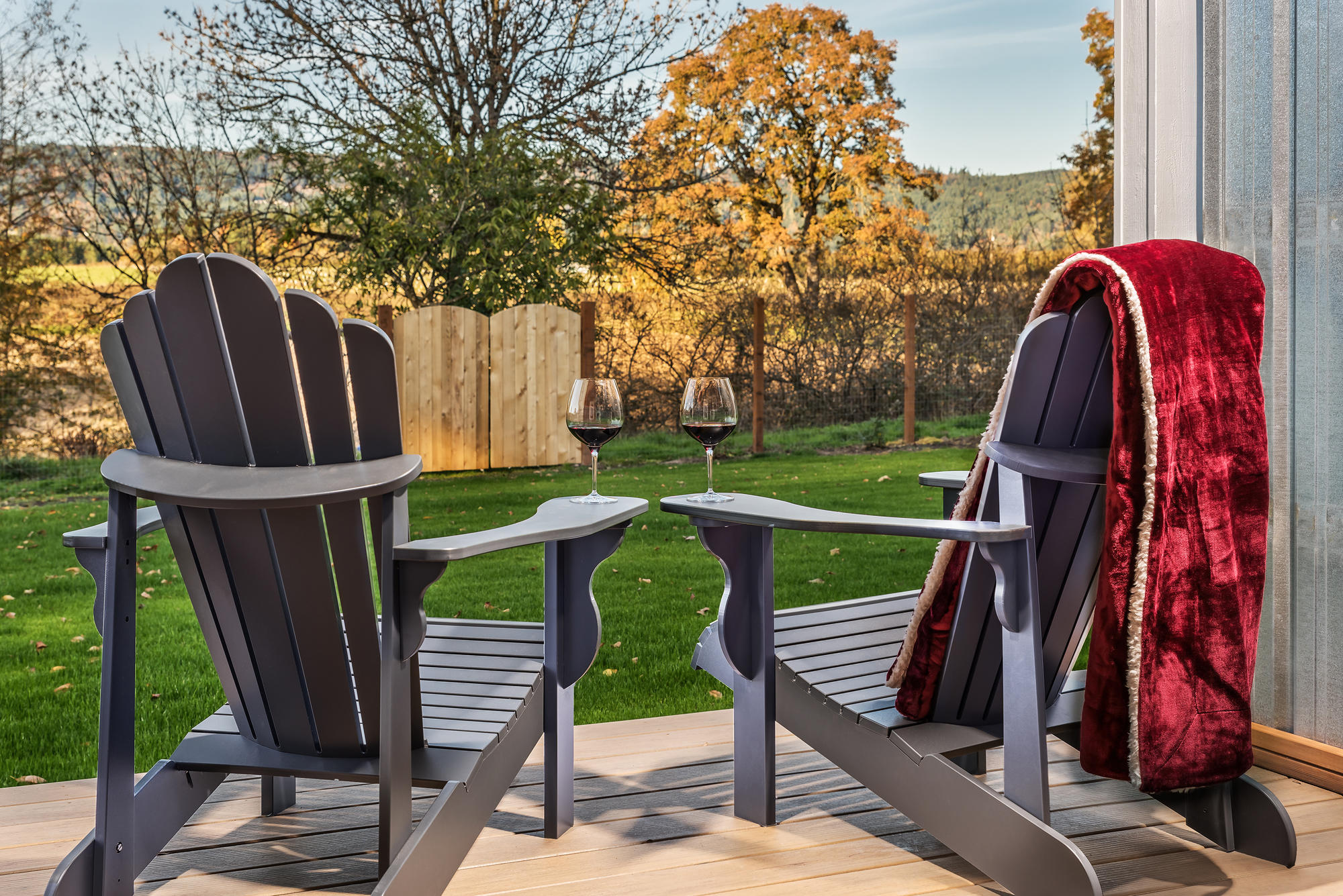 Two blue Adirondack chairs with glasses of red wine on a deck by a green lawn.