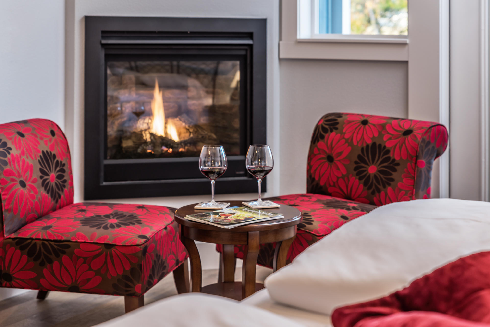 Red chairs with glasses of wine on a table by a fireplace.